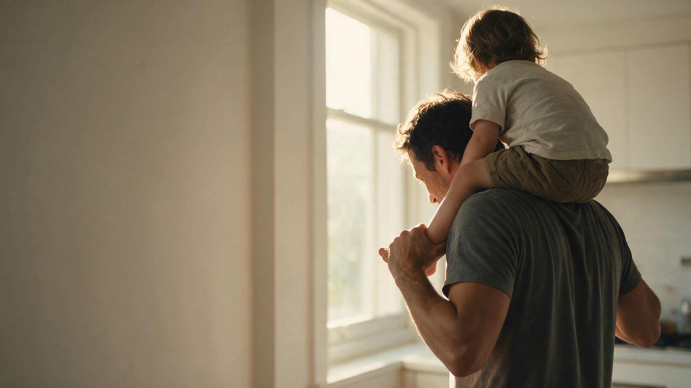 A father carrying a child on his shoulders toward a bright kitchen window