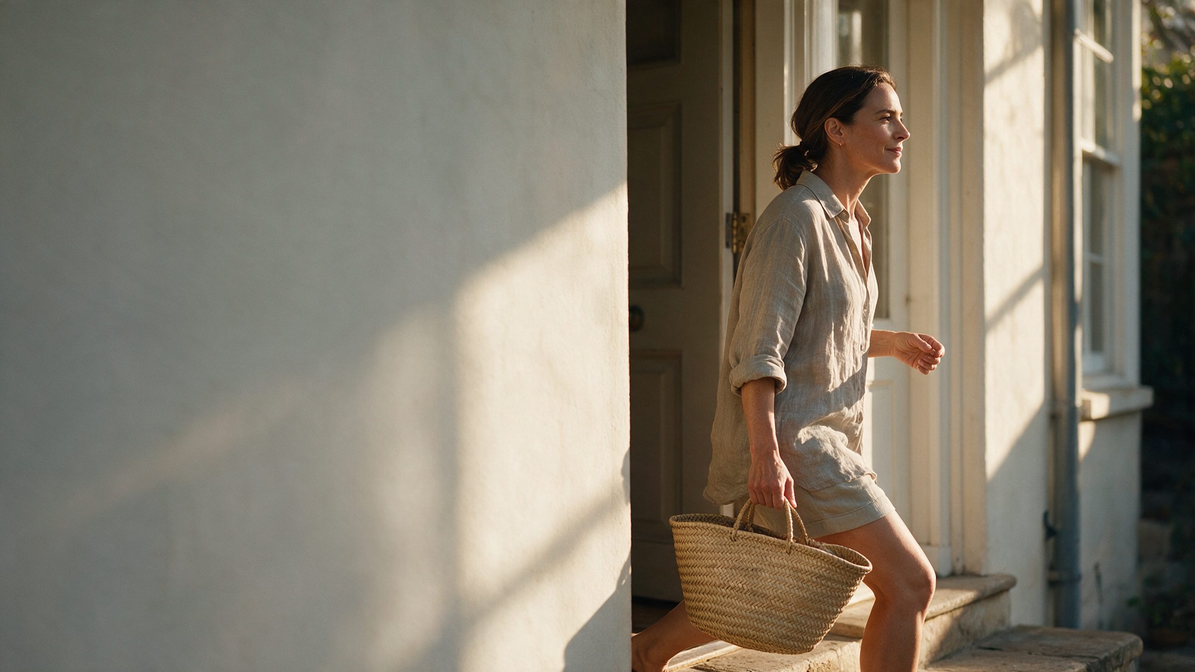 A woman stepping out of a sunlit doorway in the morning, market basket in hand