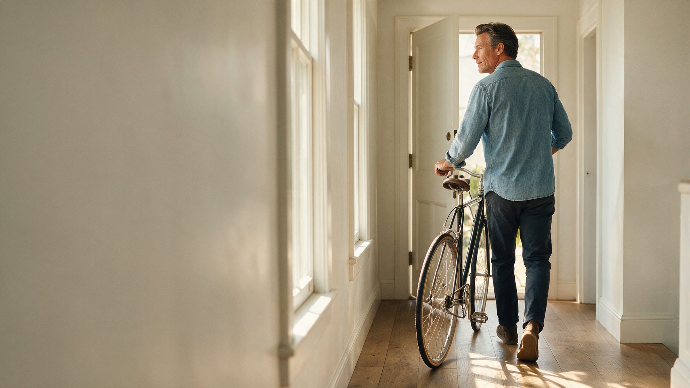 A man wheeling a bicycle through a bright hallway toward an open door